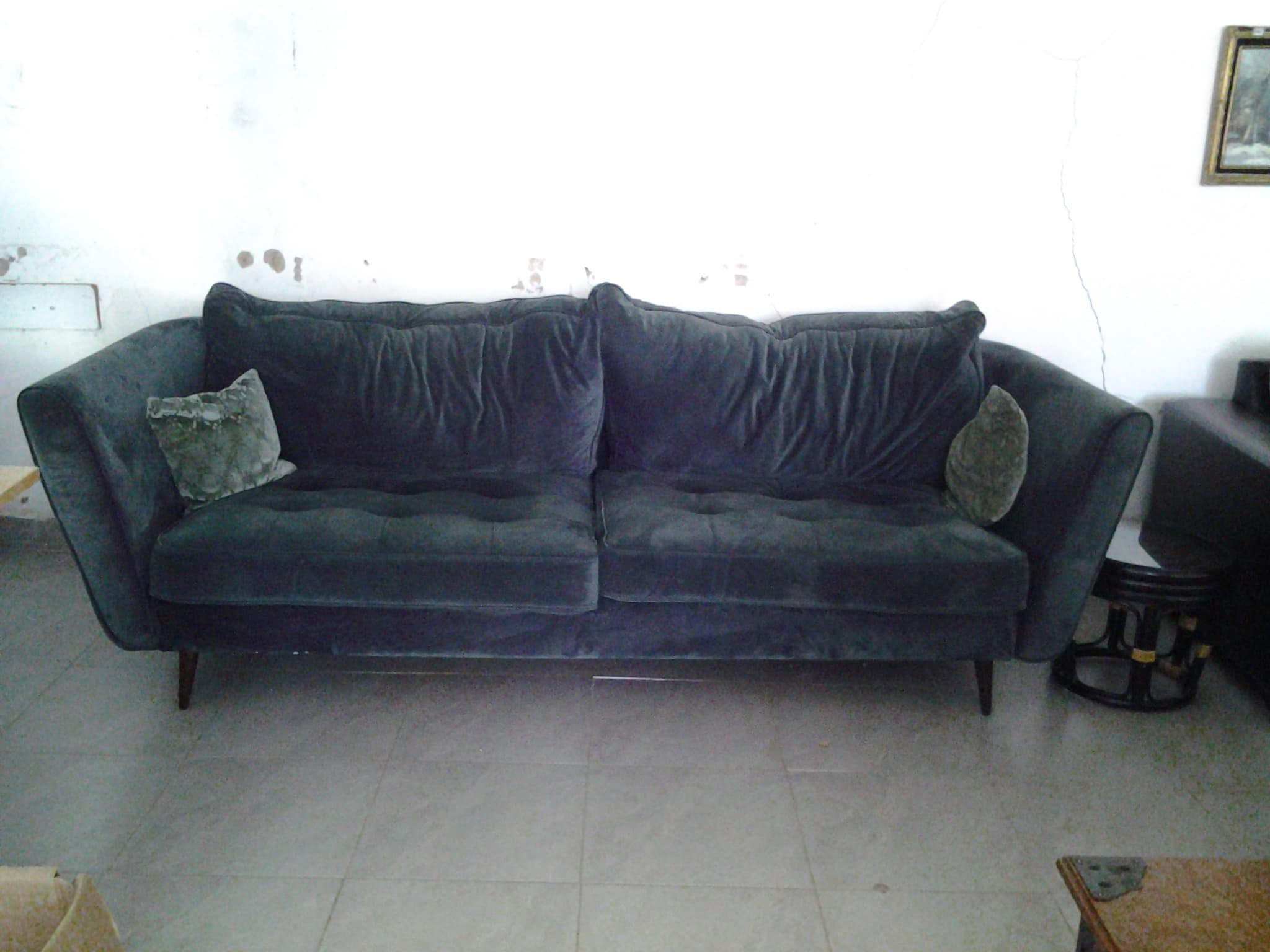 Dark blue velvet sofa with two throw pillows against a white wall on tiled flooring.